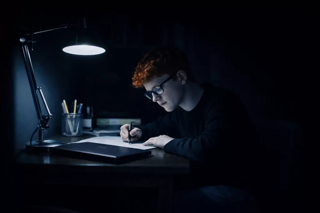 Un adolescent HPI seul à son bureau qui travaille avec une lampe allumée.