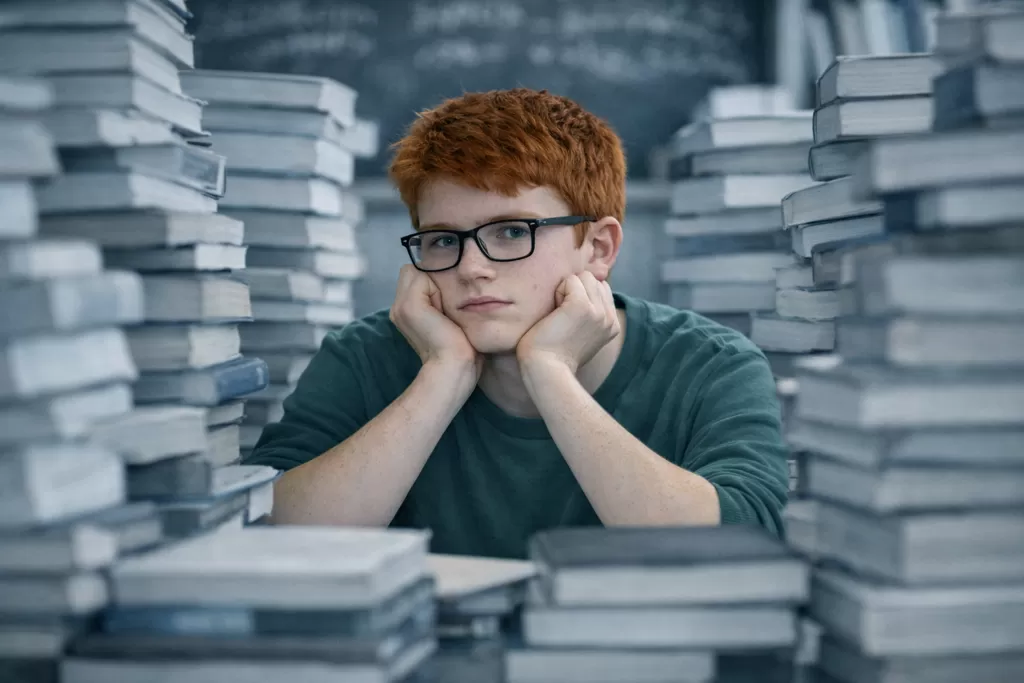 Un adolescent HPI qui se tient la tête au milieu de piles de livres.